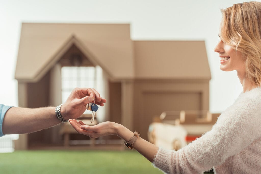A smiling woman standing outside her new house in Surrey, receiving the keys from a real estate agent, symbolizing the excitement of homeownership.